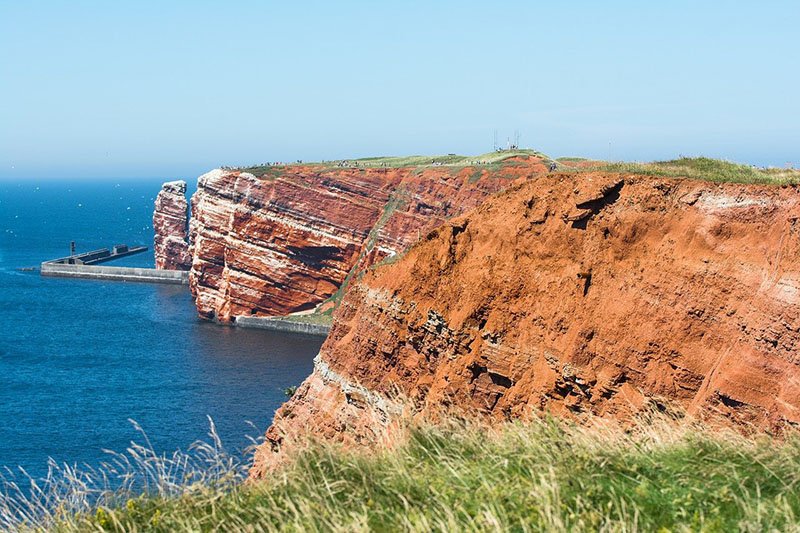 De rode klifkust van Helgoland onder een blauwe lucht, met de Lange Anna en een golfbreker op de achtergrond