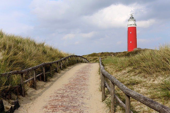 Een pad van klinkers tussen houten hekken door de duinen naar de rode vuurtoren van Texel