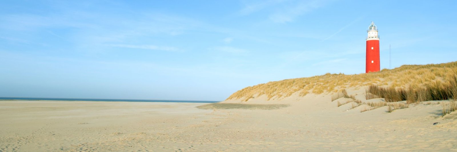 Een rode vuurtoren boven de duinen, met een leeg strand ervoor