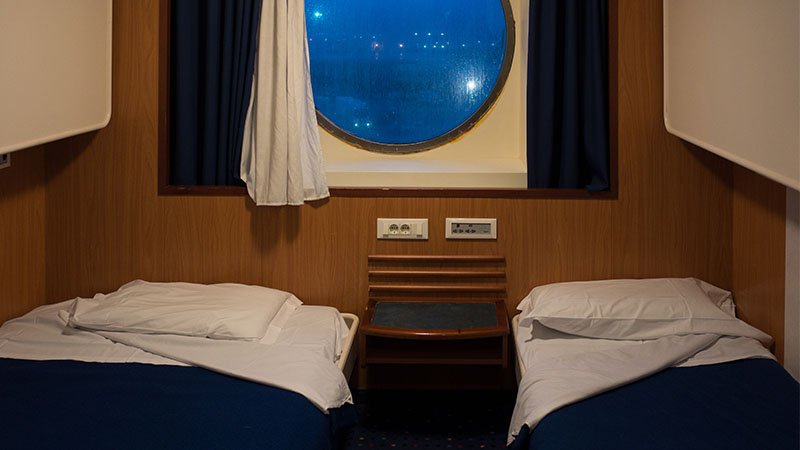 Twee opgemaakte eenpersoonsbedden met wit beddengoed en donkerblauwe spreien in een houten hut, met daartussen een klein tafeltje, twee stopcontacten en een ronde patrijspoort waarachter havenlichten branden