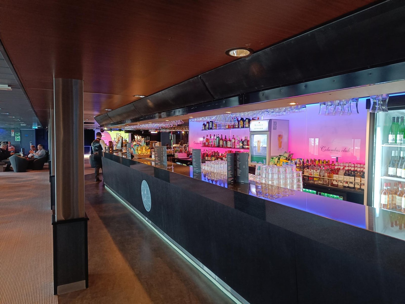 De lange bar aan boord met een verlichte flessenwand en glazen aan het plafond; op het glas erachter staat Columbus Club