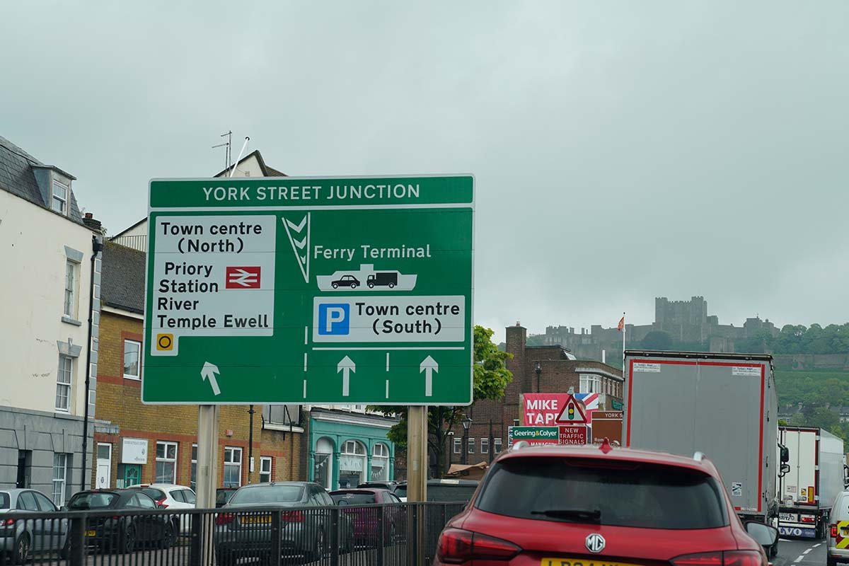 Een groot groen wegwijzerbord boven de weg in Dover met York Street Junction, Town centre North, Priory Station en Ferry Terminal, met daarachter Dover Castle op de heuvel en een rode auto en vrachtwagens in de file