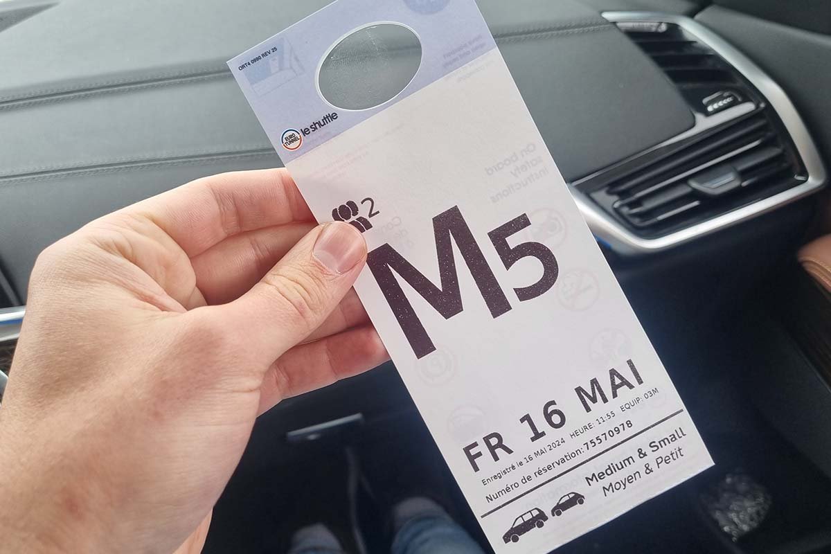 Hand met een LeShuttle-spiegelkaart in de auto: een grote letter en cijfer, de vertrekdag en rechtsonder de categorie Medium & Small met twee autootjes
