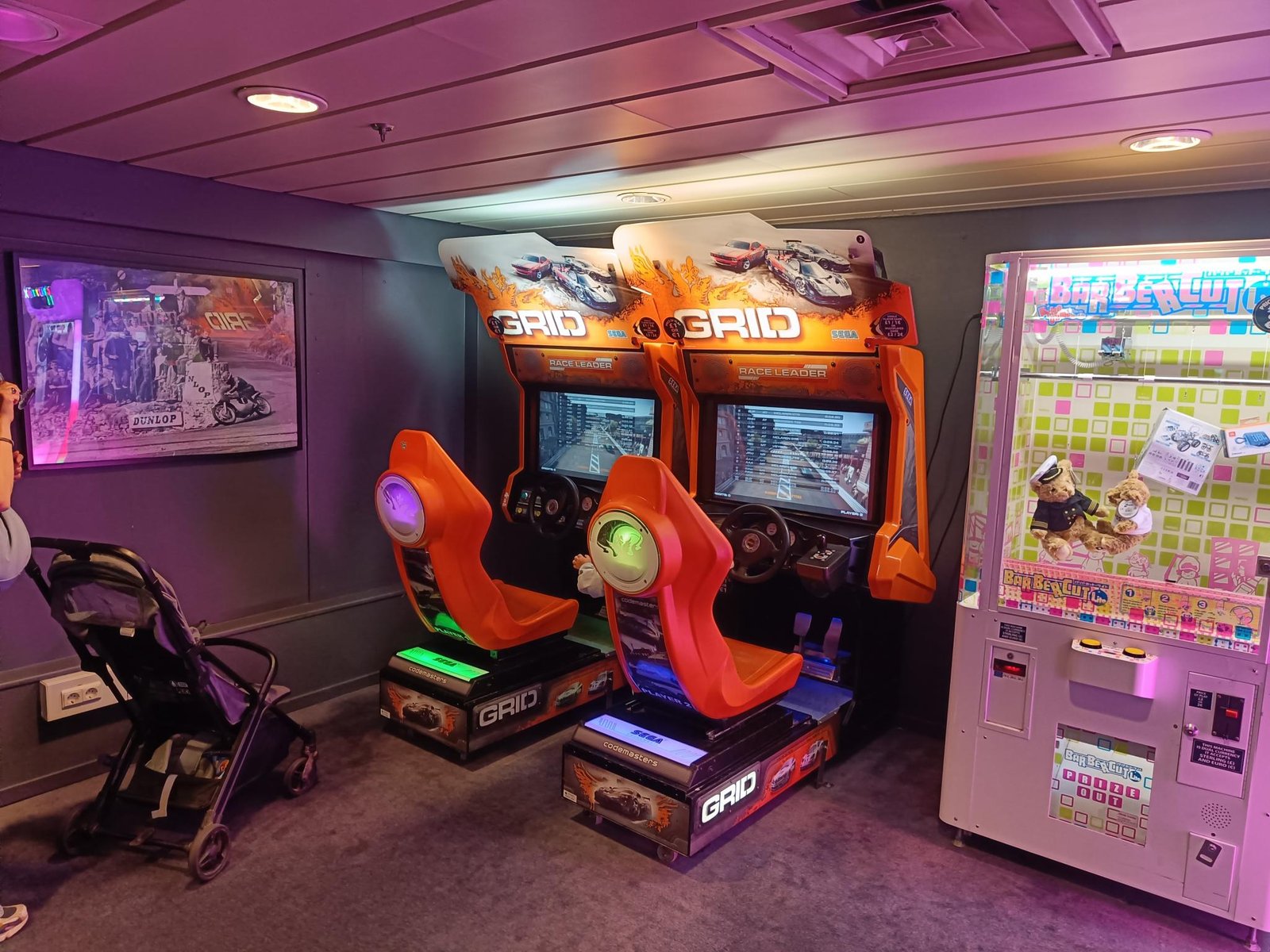 Twee oranje racegamekasten en een grijpmachine in de speelhal aan boord, met een kinderwagen ernaast