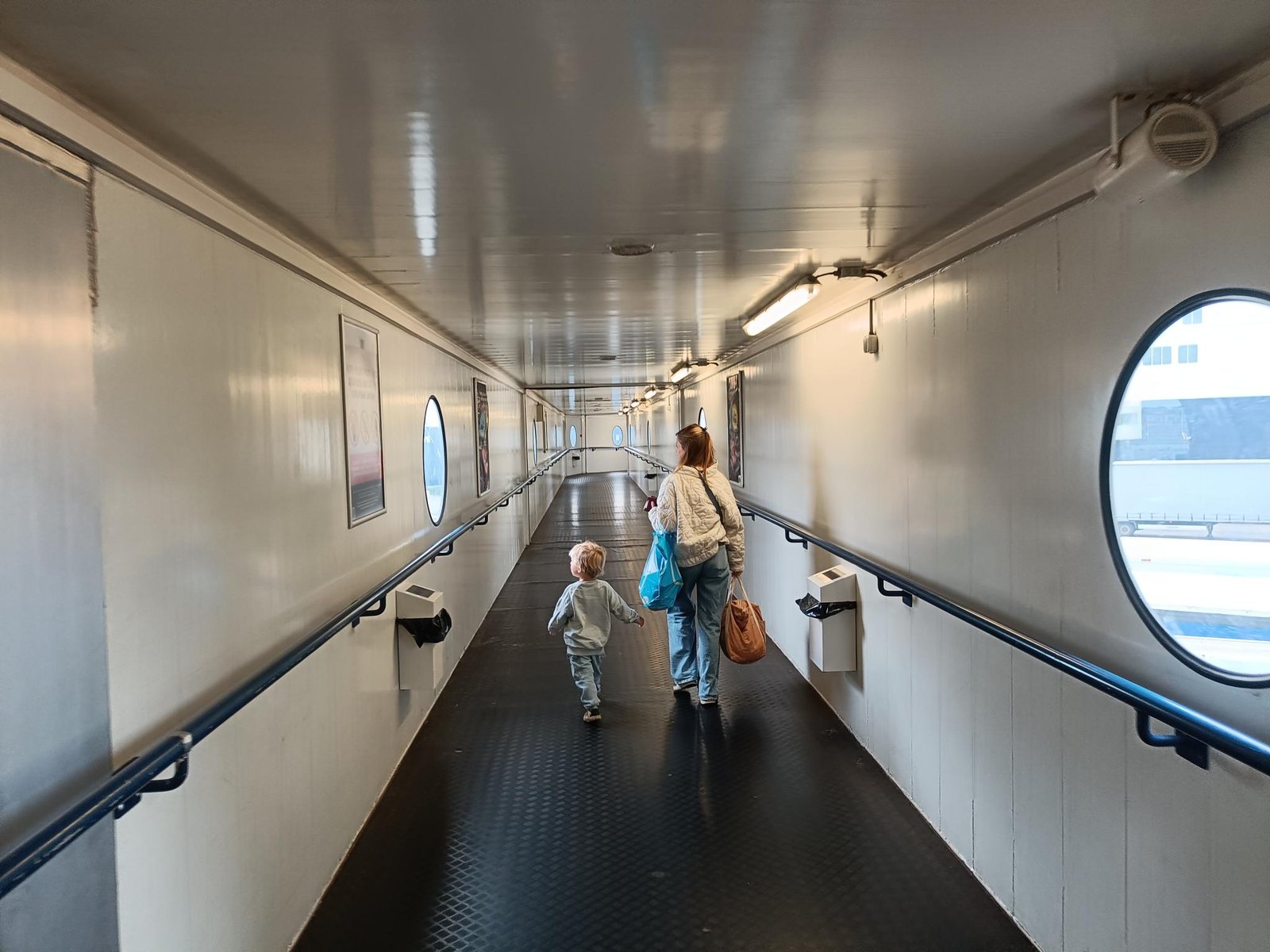 Een volwassene met tassen en een peuter lopen door de overdekte loopbrug van de terminal naar het schip