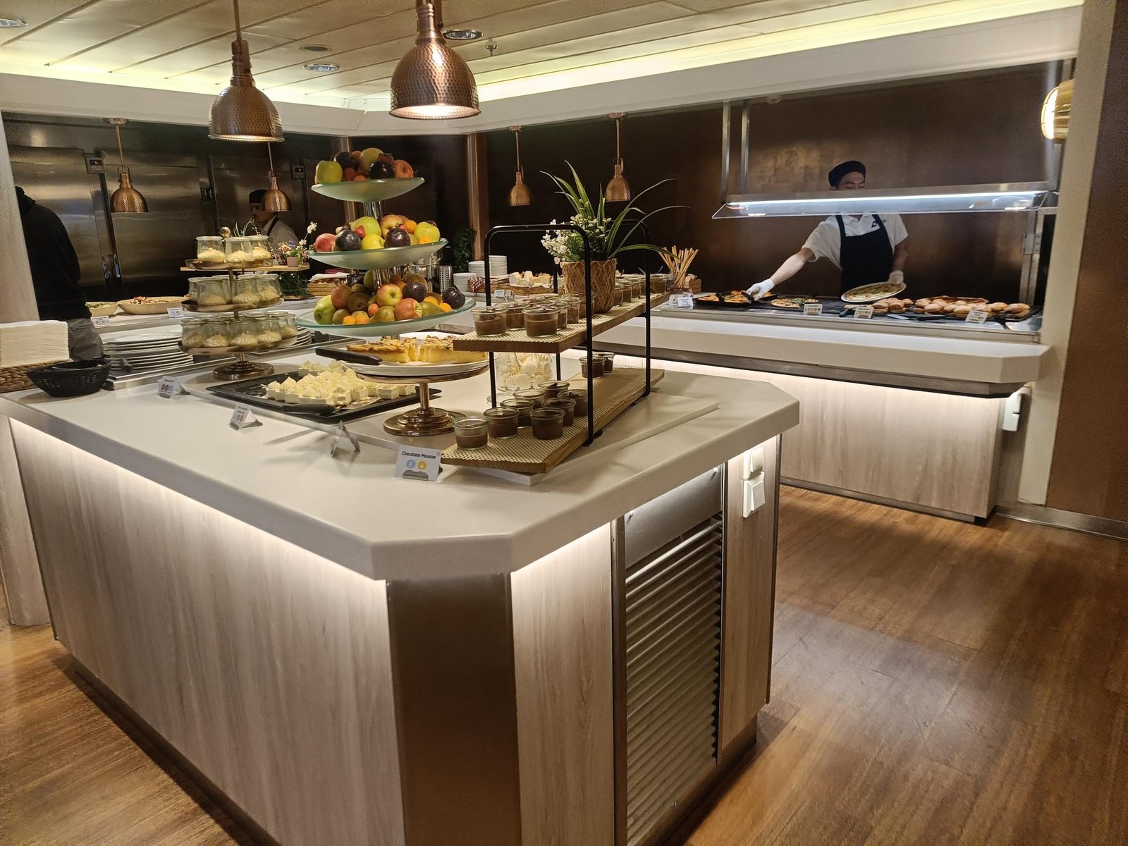 Het buffeteiland met desserts en fruit in het buffetrestaurant, met erachter een kok aan de warme balie