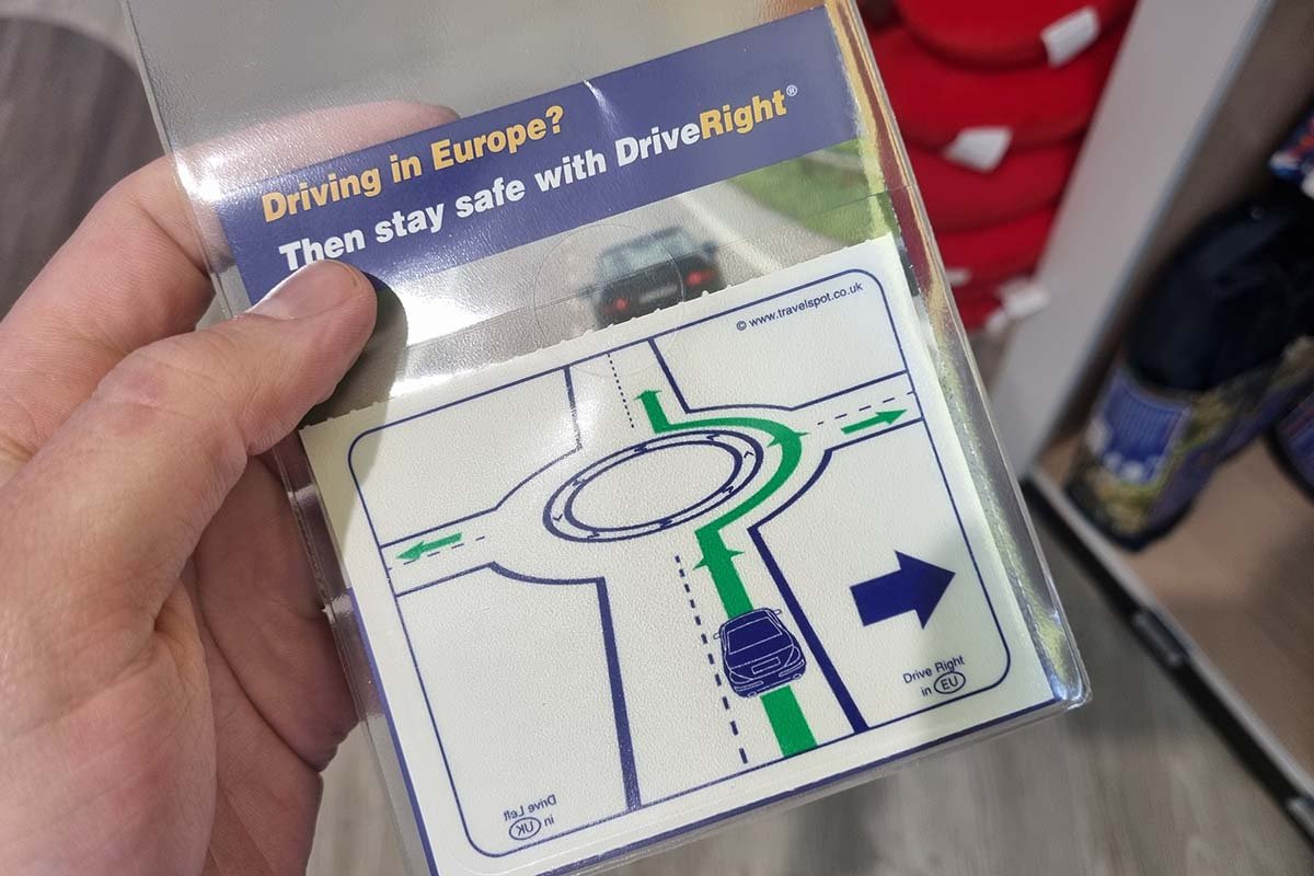 Een hand houdt een verpakt raamstickertje vast met de tekst Driving in Europe, then stay safe with DriveRight en een tekening van een rotonde met een groene lijn die de te volgen richting aangeeft