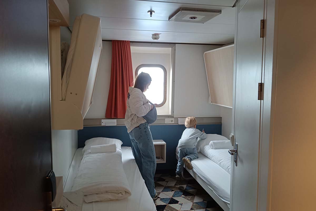 Een vierpersoonshut op een veerboot met een patrijspoort en een rood gordijn, twee opgemaakte onderbedden, twee opgeklapte bovenbedden en een klein kind dat op een van de bedden klimt