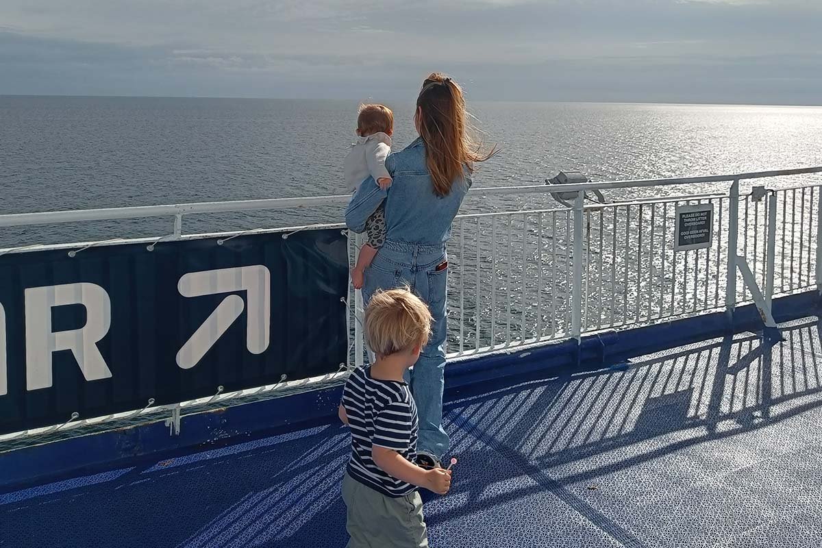 Een volwassene met een baby op de arm en een jongetje in een gestreept shirt kijken vanaf het buitendek van een veerboot over de reling uit over de zee, met de laagstaande zon op het water