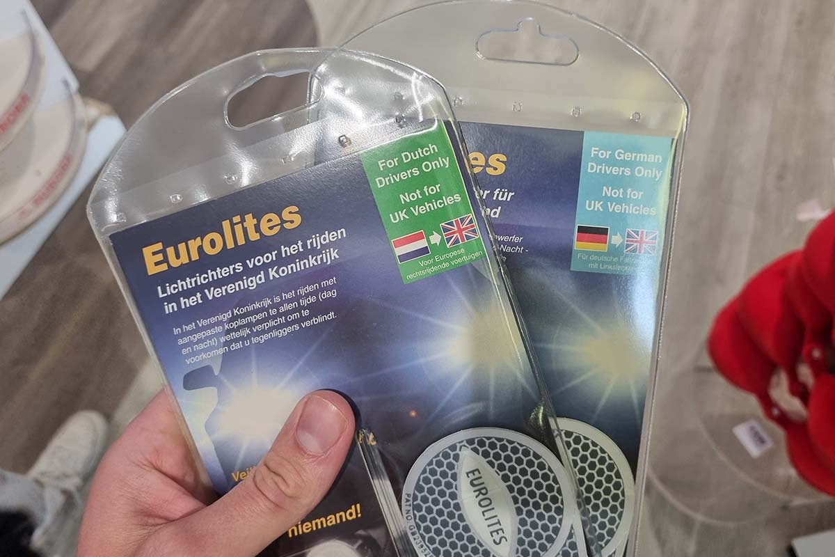 Een hand houdt twee verpakte sets lichttrichters vast, met op de linker Eurolites, lichttrichters voor het rijden in het Verenigd Koninkrijk en een groen label For Dutch Drivers Only, Not for UK Vehicles