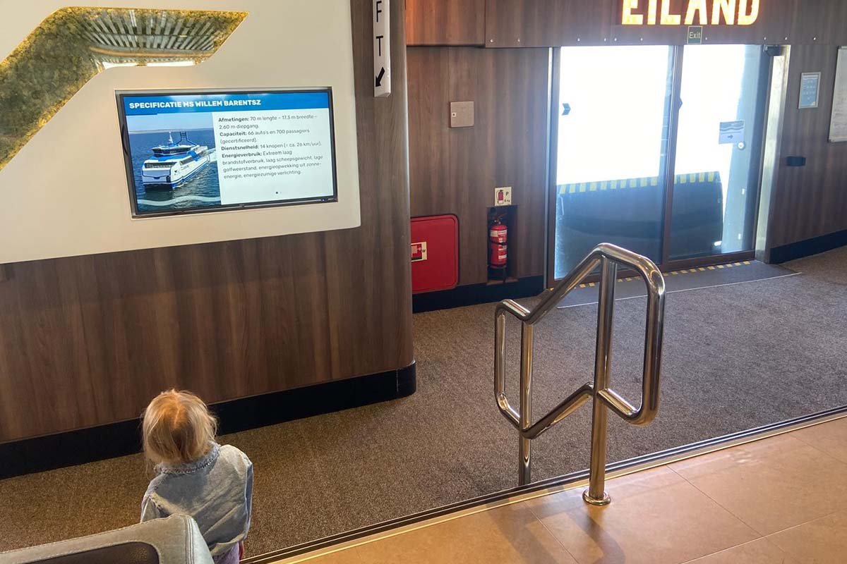 Een peuter zit in de passagiersruimte van een veerboot naar een beeldscherm te kijken waarop de specificaties van de MS Willem Barentsz staan, met afmetingen, capaciteit en dienstsnelheid