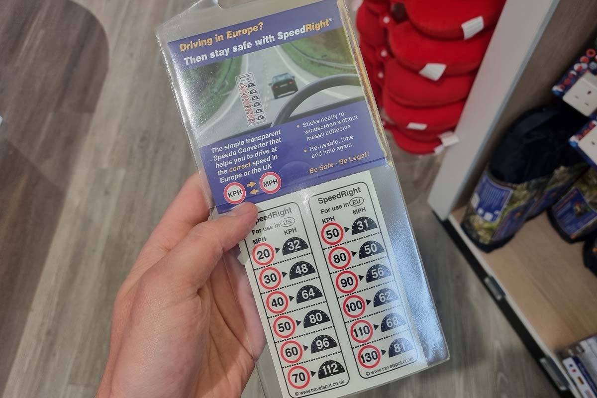 Een hand houdt een verpakte omrekensticker vast met twee kolommen snelheidsborden: links For use in UK met 20 tot 70 mijl per uur omgerekend naar kilometers, rechts For use in EU met 50 tot 130 kilometer per uur omgerekend naar mijlen