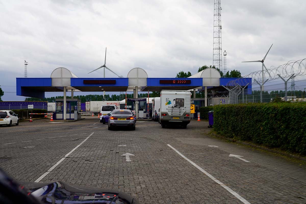 De check-in van de veerhaven: een rij cabines onder een blauwe luifel, met een auto, een bestelbus en een camper ervoor en windmolens erachter