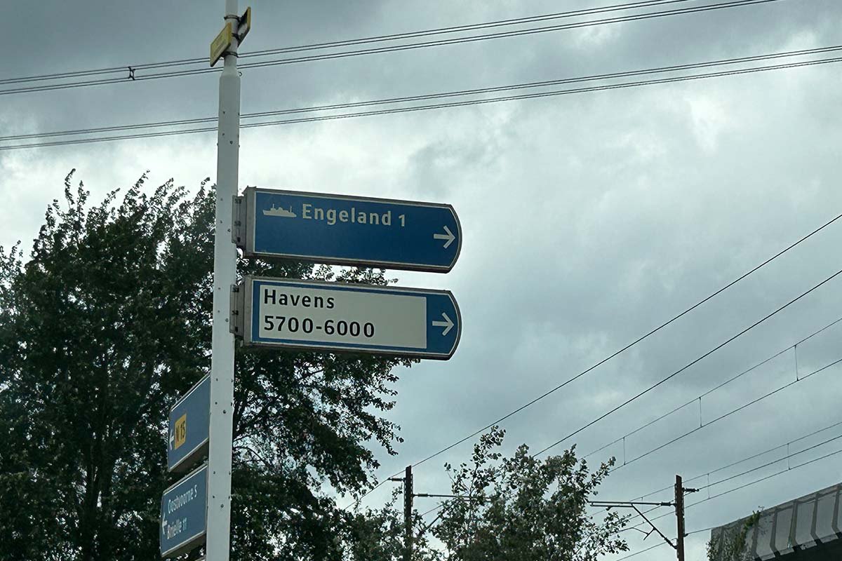 Blauw wegwijzerbord langs de weg met een scheepje en het woord Engeland, daaronder een wit bord met Havens 5700-6000, onder een bewolkte lucht met bovenleidingen