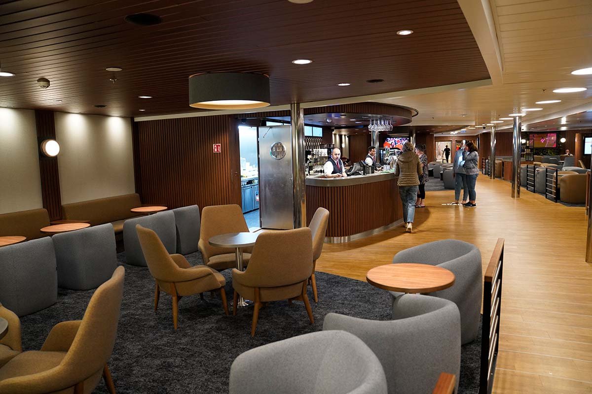 De bar aan boord met een gebogen toog, een barman erachter en fauteuils eromheen