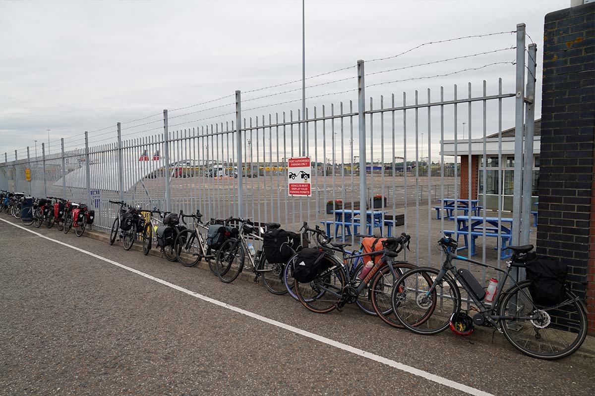 Een rij volgepakte fietsen met bagagetassen tegen een stalen hek bij de terminal van Harwich, met picknicktafels erachter