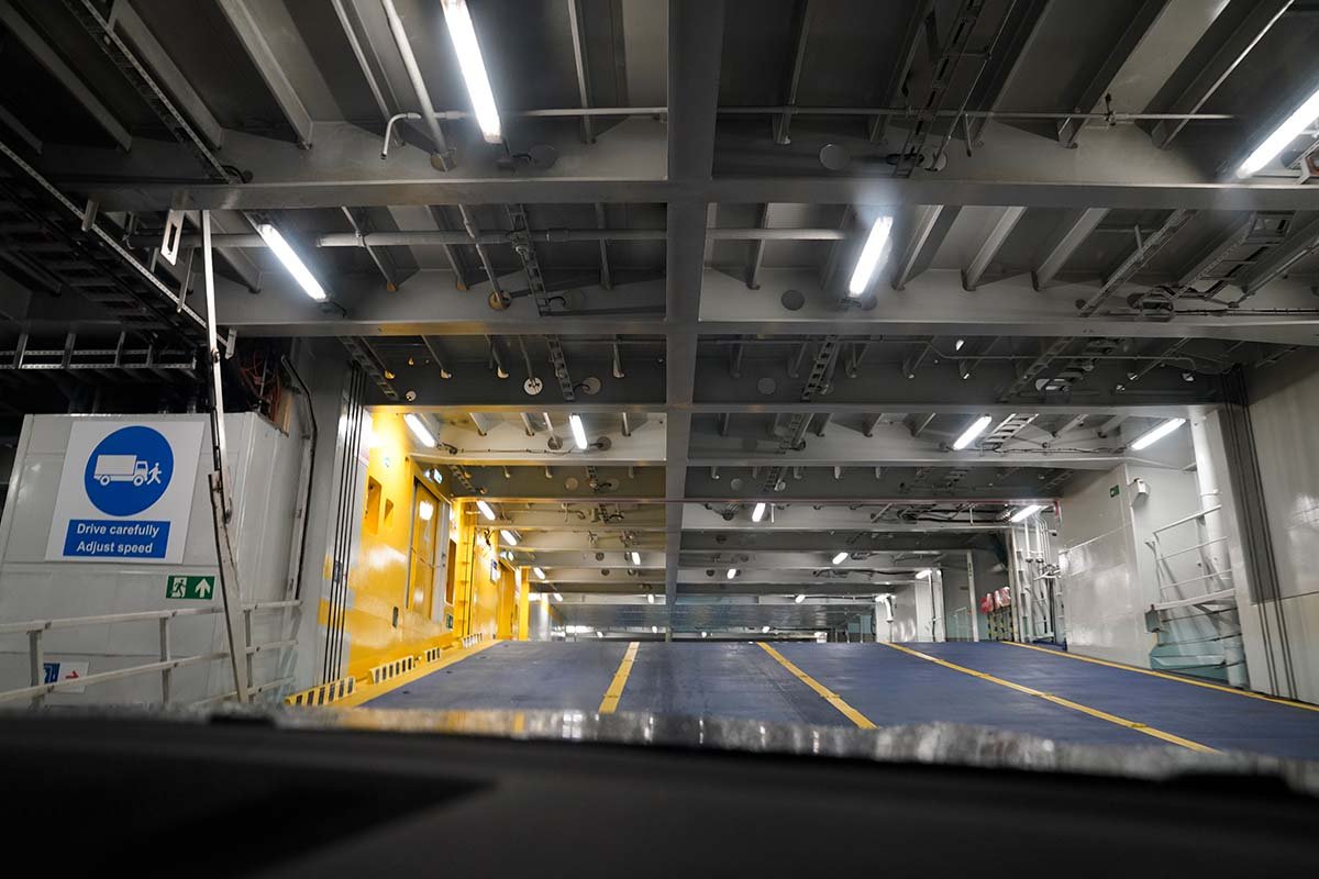 Het lege autodek van de veerboot, gezien vanachter de voorruit, met gele rijlijnen en een bord Drive carefully, adjust speed