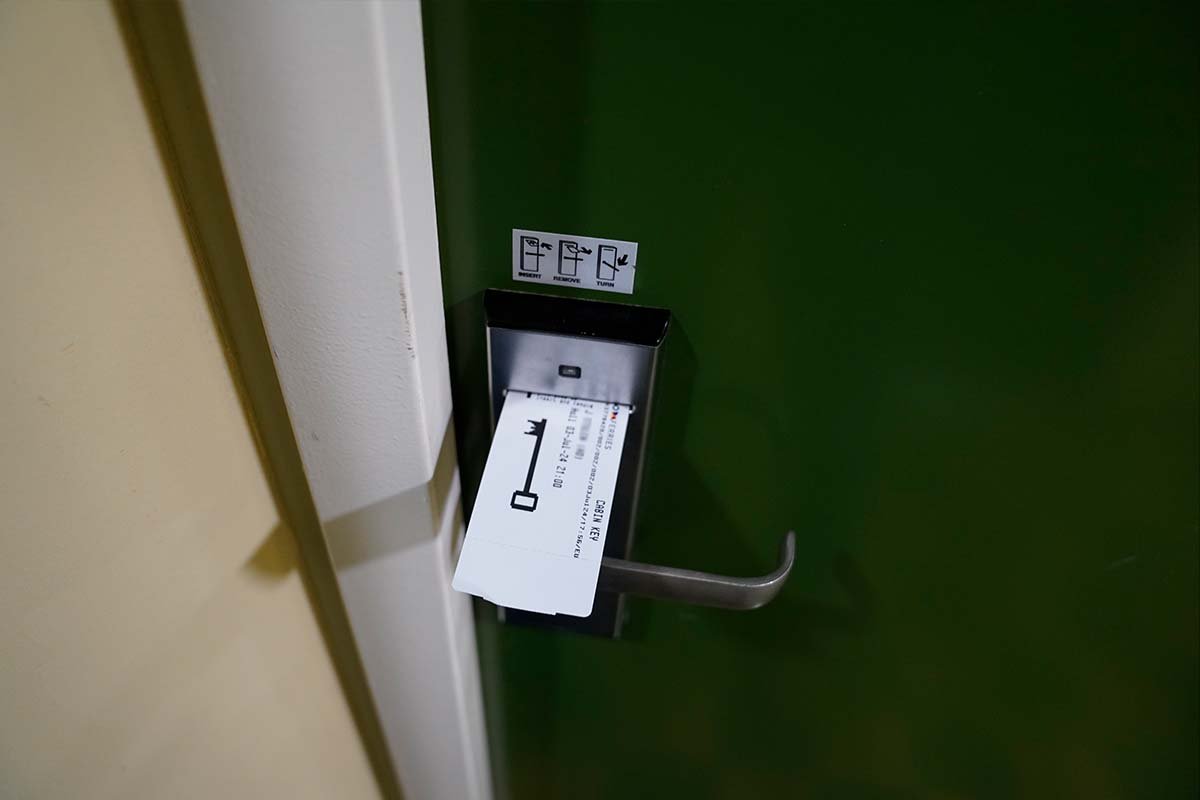 Het papieren kaartje steekt in de gleuf van het slot naast de klink van een donkergroene hutdeur; erboven zit een sticker met de stappen insert, remove en turn