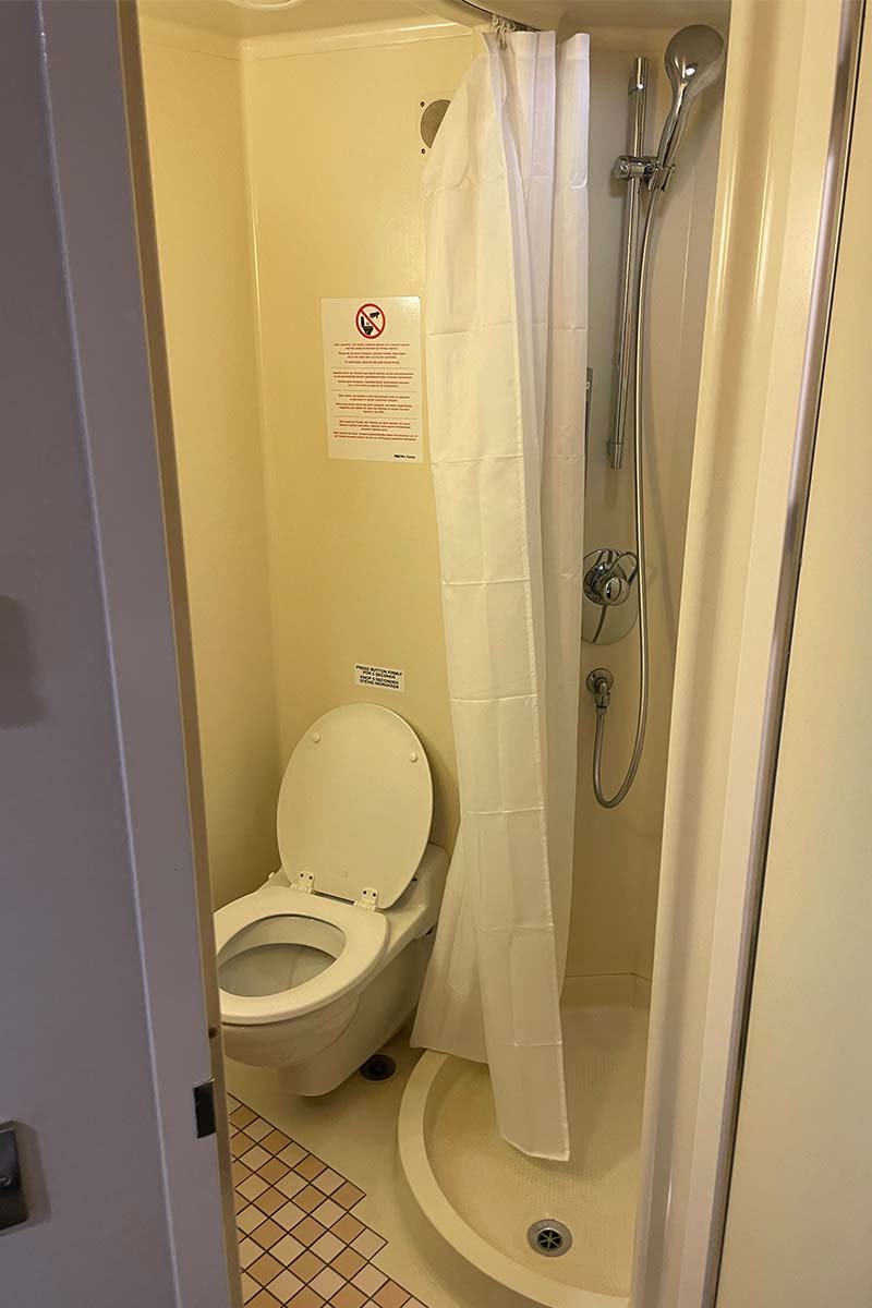 De kleine badkamer van de hut, met een hangend toilet, een ronde douchebak met een wit douchegordijn en een handdouche, alles in crèmekleurig kunststof