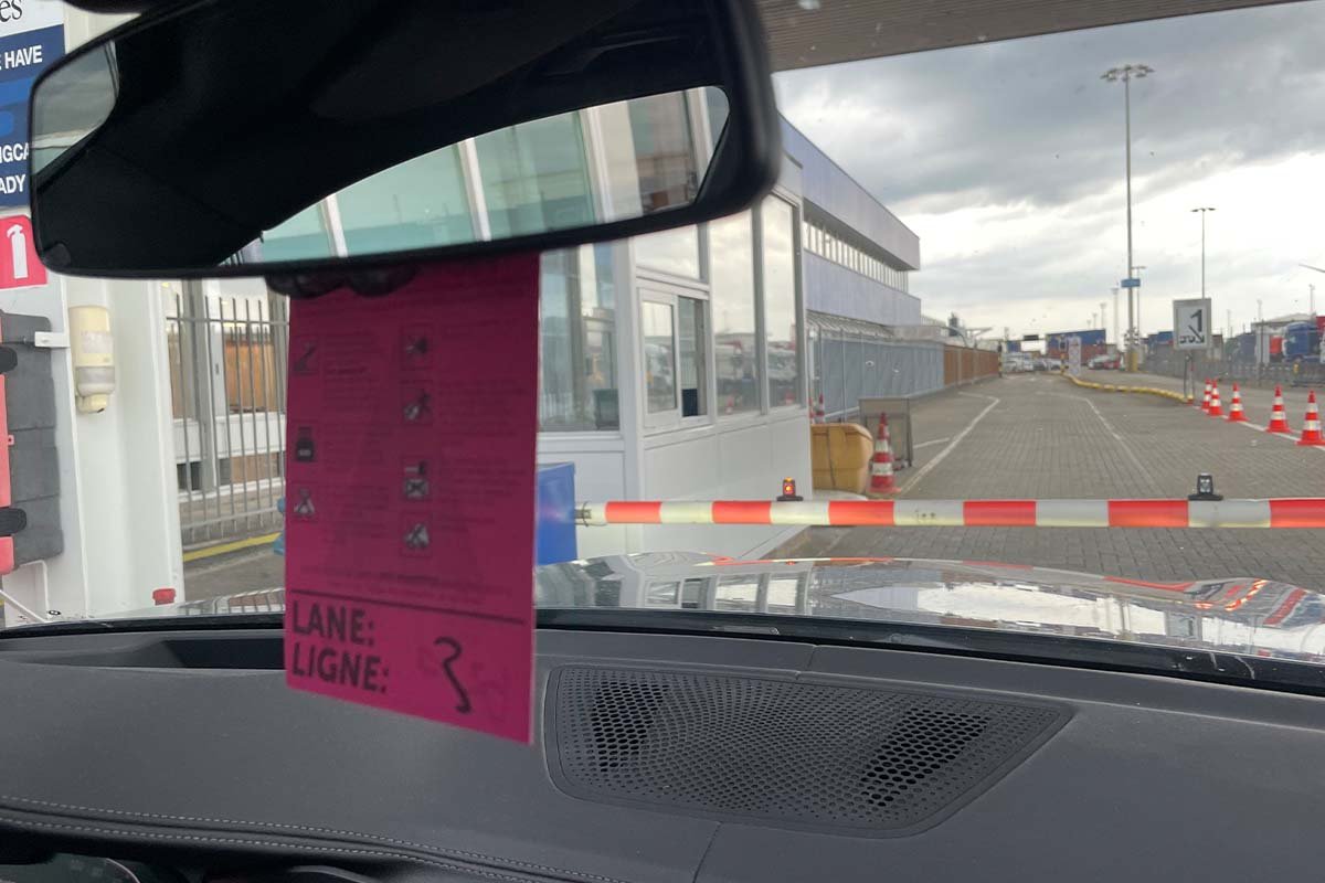 Gezicht door de voorruit van de auto op een gesloten rood-witte slagboom en een klein terminalgebouw, met aan de binnenspiegel een roze kaartje waarop Lane staat met een handgeschreven 3