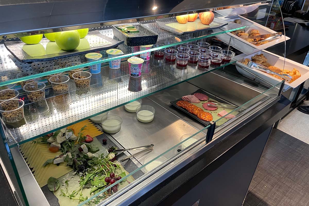 Gekoelde vitrine met appels, bakjes yoghurt en muesli, glaasjes sap, een schaal vleeswaren, croissants en salade