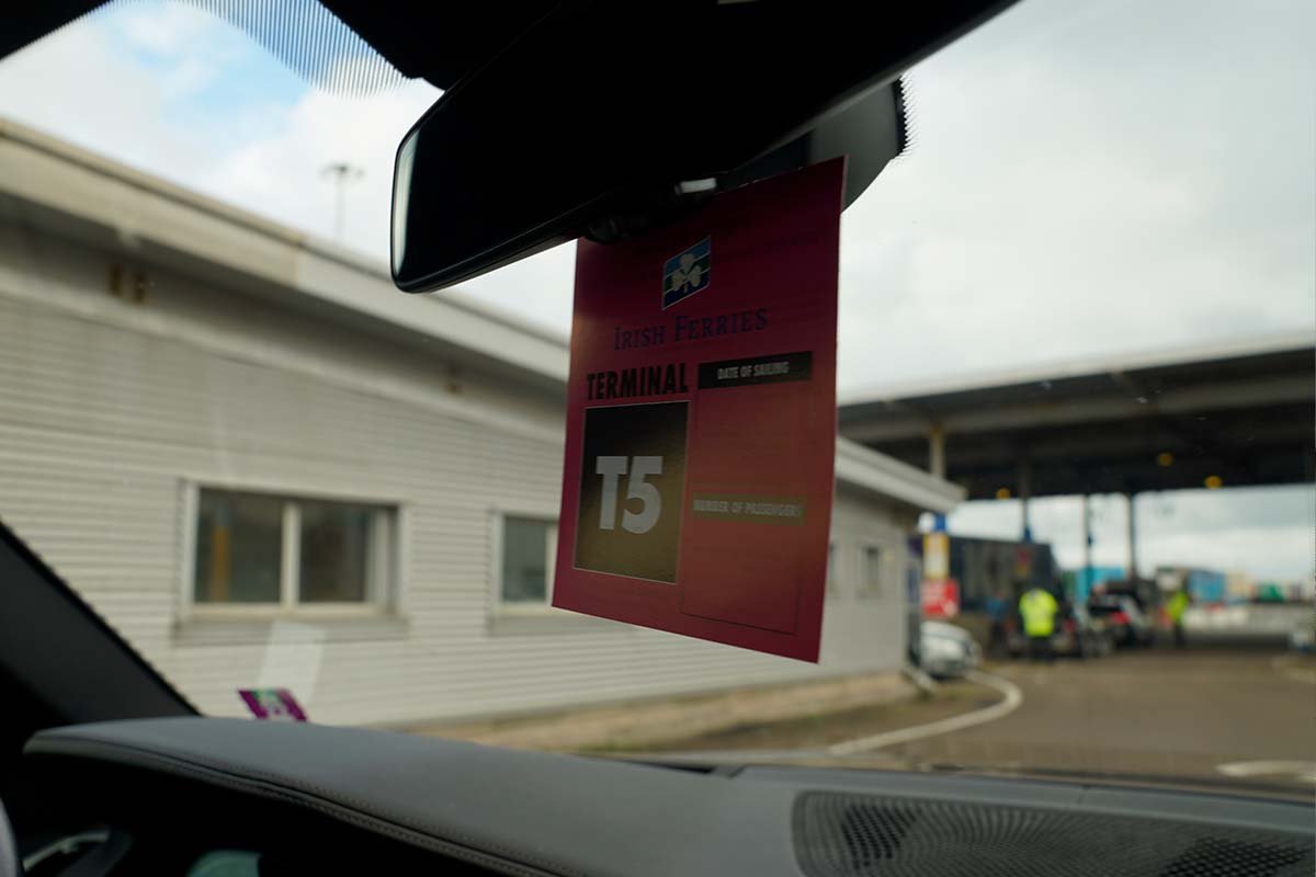 Rode kaart van Irish Ferries met de tekst Terminal T5 aan de binnenspiegel van een auto; door de voorruit de hokjes van de check-in en een medewerker in een geel hesje