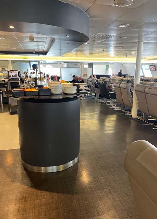 Rond zwart buffeteiland met stapels borden en schalen, bakjes bestek en fruit, en daarachter sapautomaten en rijen beige leunstoelen met passagiers