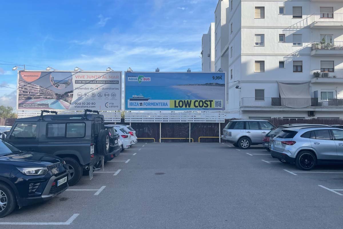 Parkeerterrein met auto's en daarachter twee reclameborden, waarvan het rechter een veerboot naar Formentera aanprijst met een bedrag van 9,99 euro
