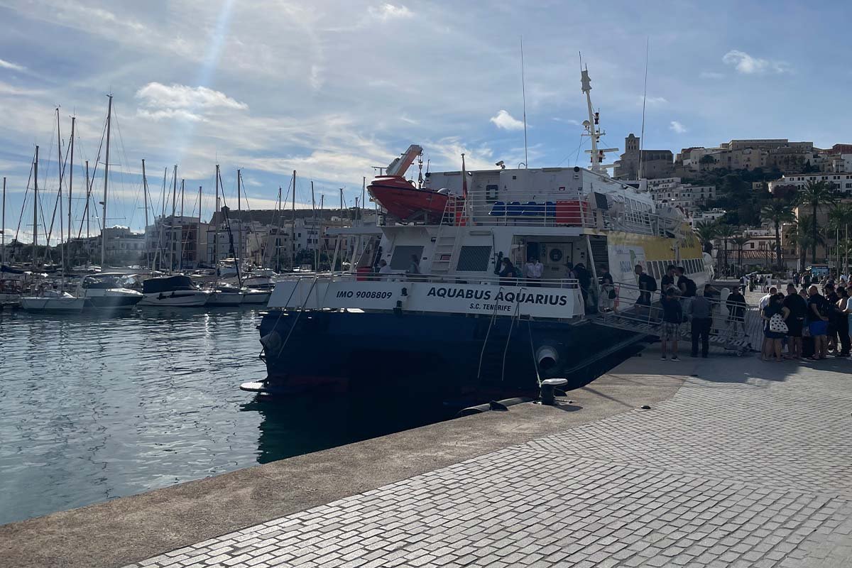 Blauw-witte snelle veerboot langs de kade in de haven van Ibiza met een rij wachtende passagiers ernaast, en op de heuvel erachter de oude bovenstad