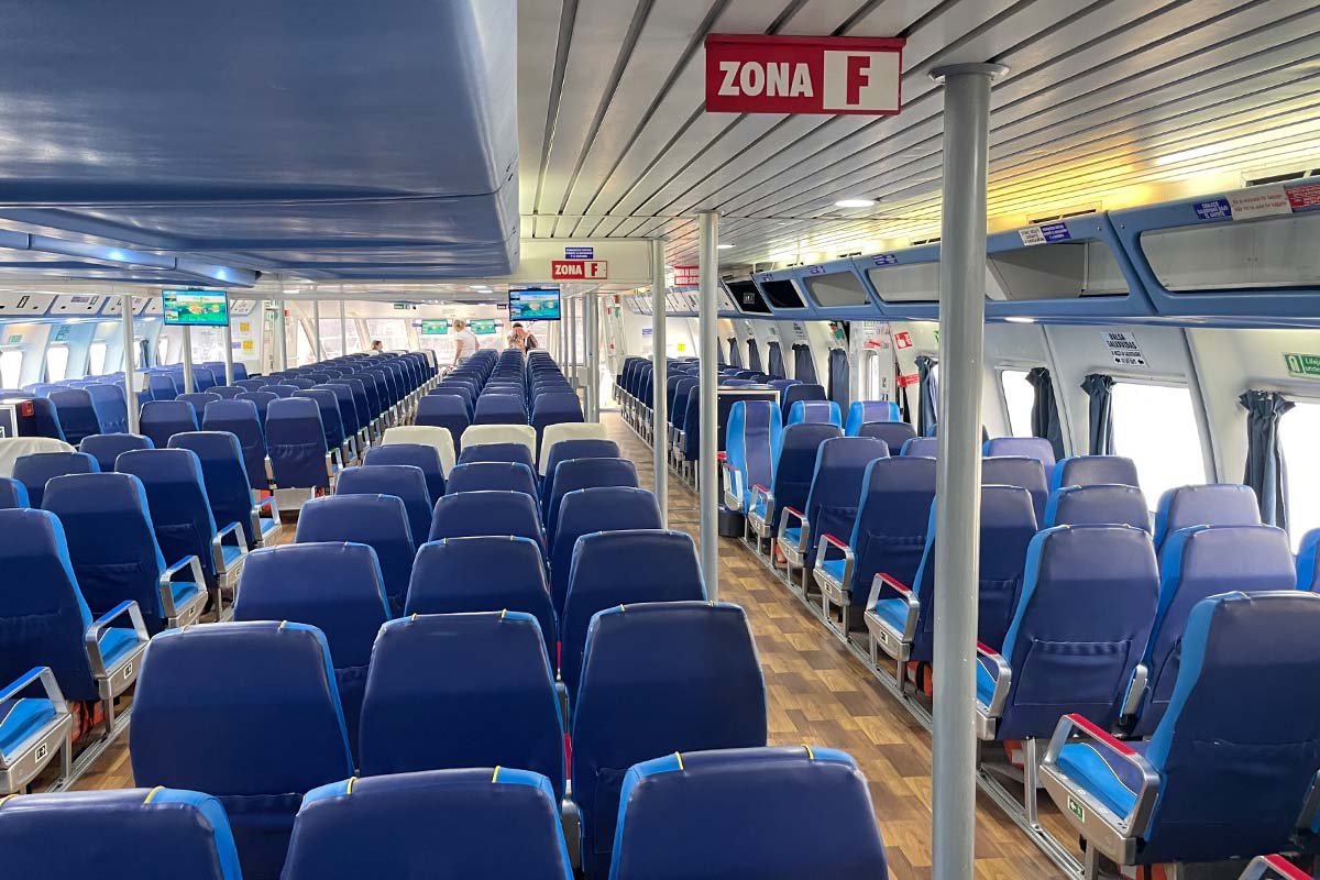 Passagiersdek van een snelle veerboot met lange rijen blauwe stoelen, een vloer met houtlook en een rood bordje met Zona F aan het plafond