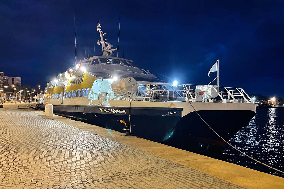 Dezelfde snelle veerboot 's avonds afgemeerd aan een verlichte kade, met dekverlichting aan onder een donkerblauwe lucht
