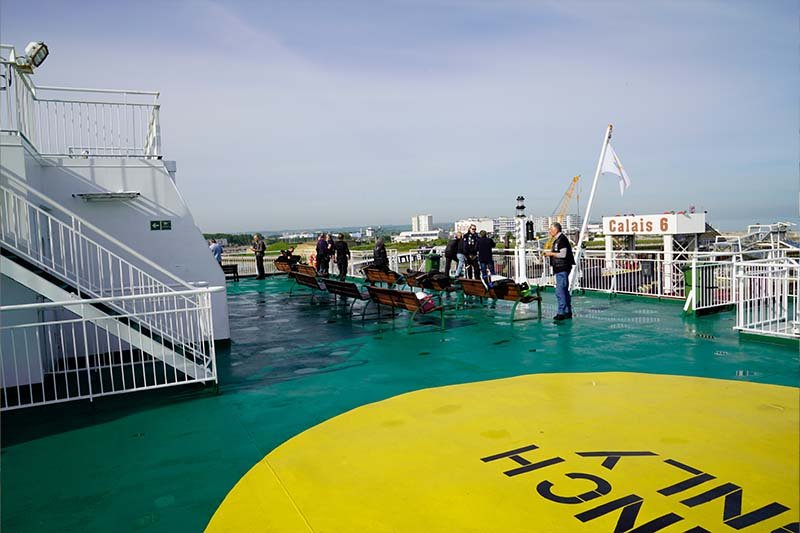 Buitendek met houten bankjes en mensen aan de reling, een gele cirkel op de dekvloer en op de kade het bord Calais 6