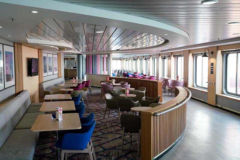 Salon aan boord van DFDS met blauwe en roze fauteuils, houten tafels en een bank langs de wand onder de ramen