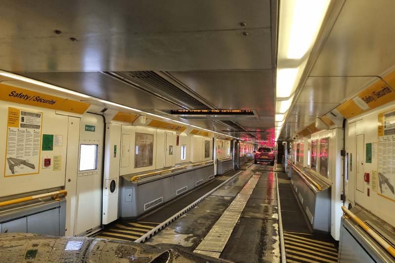 Blik door de wagon van de autotrein: een smal looppad langs de zijkant, gele veiligheidsborden en verderop een auto met rode achterlichten