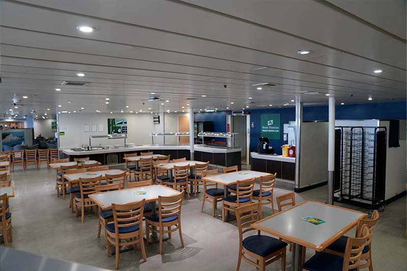 Zelfbedieningsrestaurant van Irish Ferries met lege houten stoelen met blauwe zittingen, witte tafels en een buffetlijn op de achtergrond