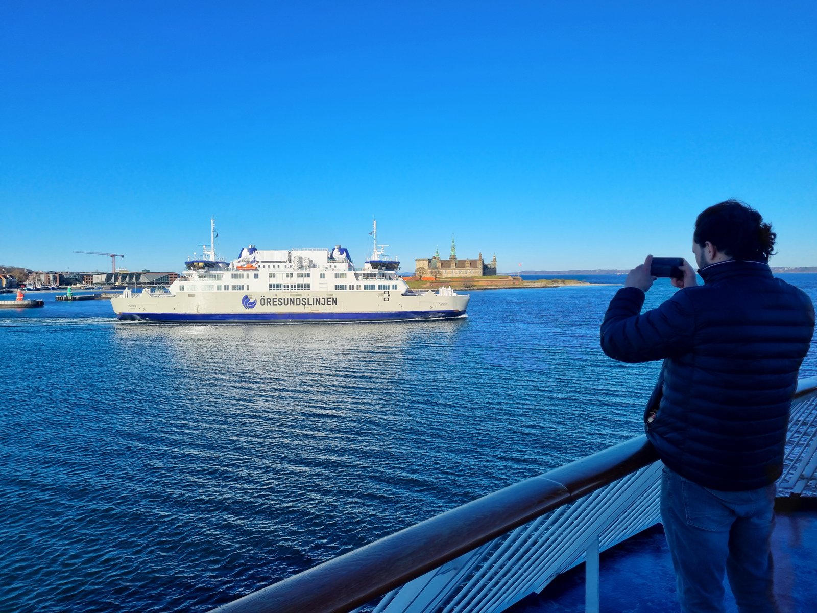 Vanaf het dek van een veerboot fotografeert een man de witte Aurora van Öresundslinjen die voorbij vaart, met kasteel Kronborg op de achtergrond onder een strakblauwe lucht