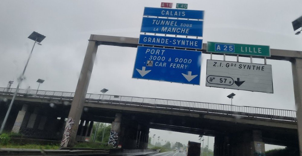 Portaal boven de A16 met blauwe borden naar Calais, Tunnel sous la Manche en Port Car Ferry, gezien door een natte voorruit