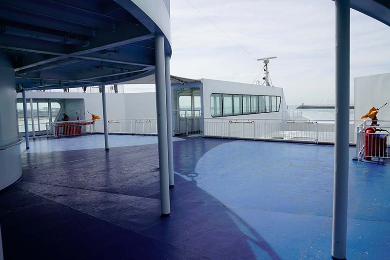 Leeg buitendek met blauwe vloer, witte palen en hekken en uitzicht op de havenmond, zonder een enkele bank of stoel