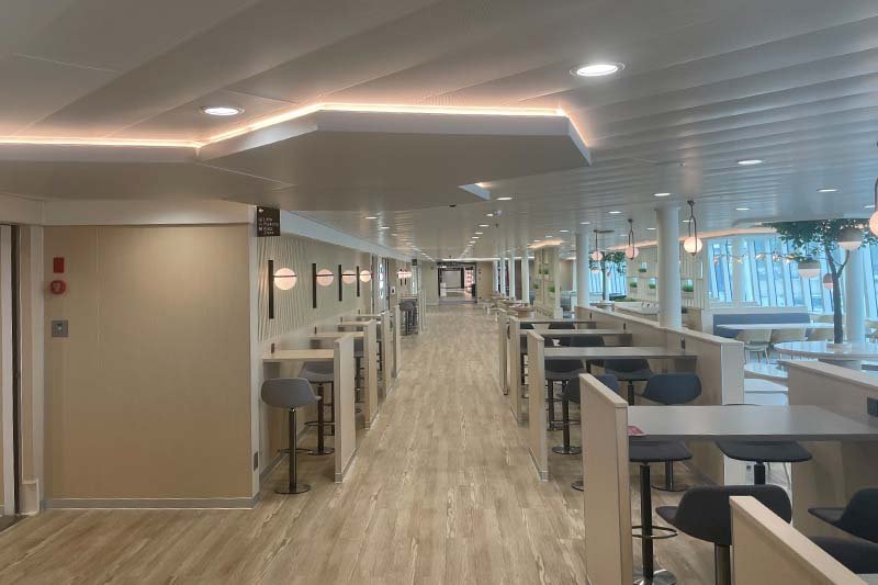 Dek aan boord van P&O Ferries met een lichte houten vloer, eettafels met barkrukken in nissen en ramen aan de rechterkant