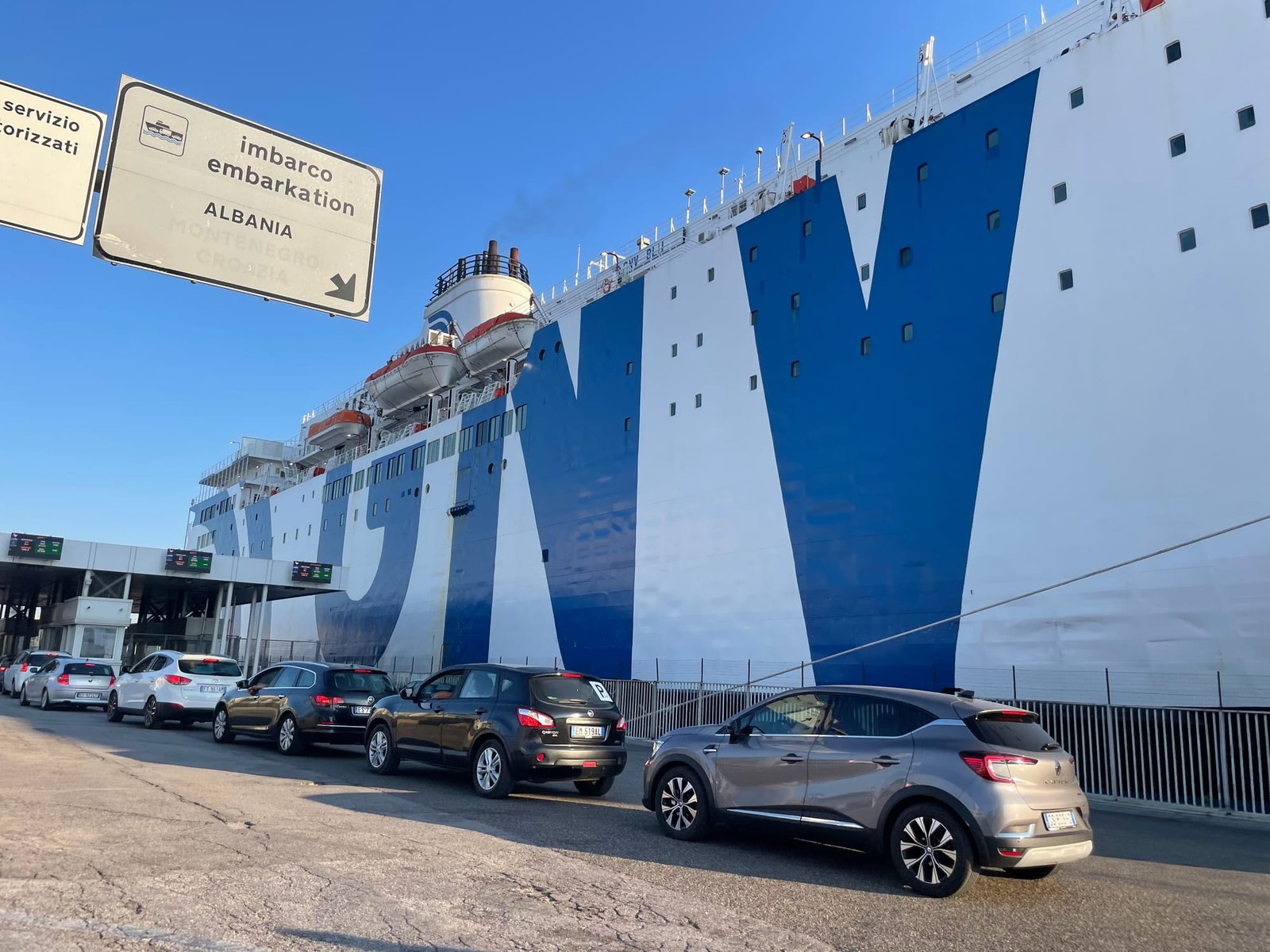 Auto's in de rij voor het inschepen in de haven van Bari, onder een bord met imbarco embarkation Albania, naast een groot wit-blauw schip van GNV