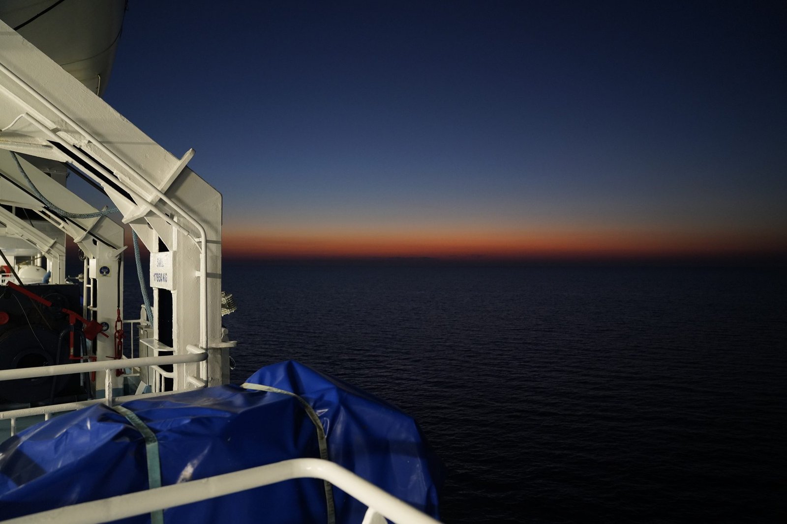 Uitzicht vanaf het dek over een donkere zee met een oranje band aan de horizon na zonsondergang, links een reddingsboot