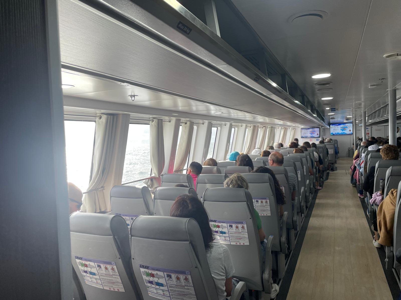 Rijen stoelen in de passagierssalon van een SNAV-draagvleugelboot, met passagiers en ramen met gordijntjes
