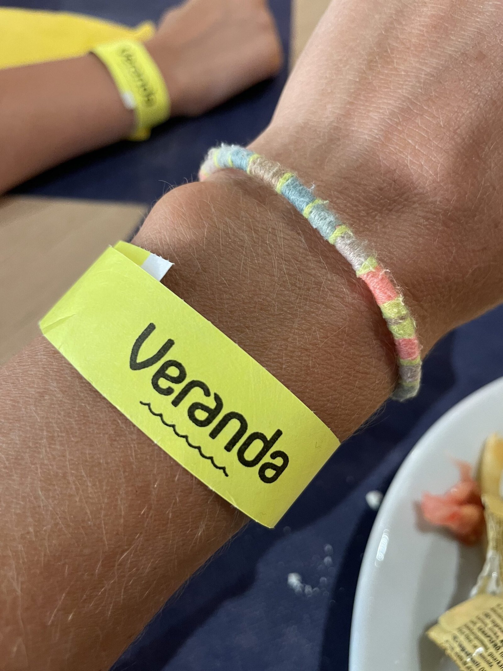 Fel geel papieren polsbandje met het woord Veranda in handschriftletters en een golflijntje eronder, om een pols naast een gevlochten vriendschapsarmbandje, met op de achtergrond een tweede arm met hetzelfde bandje en een bord eten op een donkerblauw tafelblad