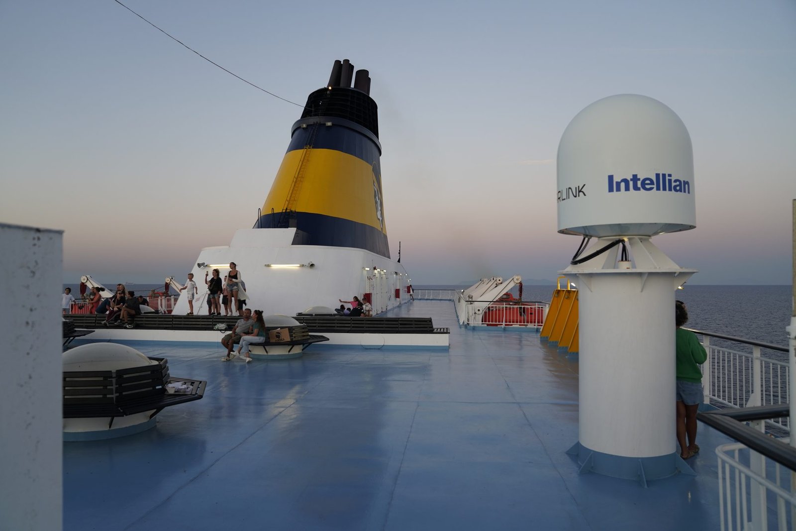 Bovendek van de veerboot in de schemering met de gele schoorsteen, mensen op banken en een satellietkoepel rechts