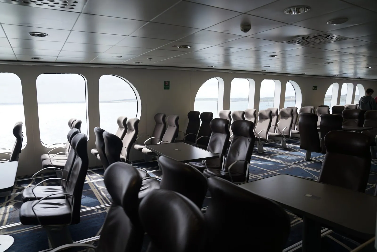 Rijen donkere leunstoelen met verstelbare rugleuning in een passagiersruimte van een veerboot, langs een wand met hoge boogramen waarachter de zee en een vlakke kustlijn te zien zijn