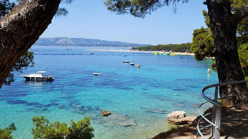 Helder turquoise water in een baai, tussen de dennen door gezien, met bootjes voor anker en in de verte een lang strand