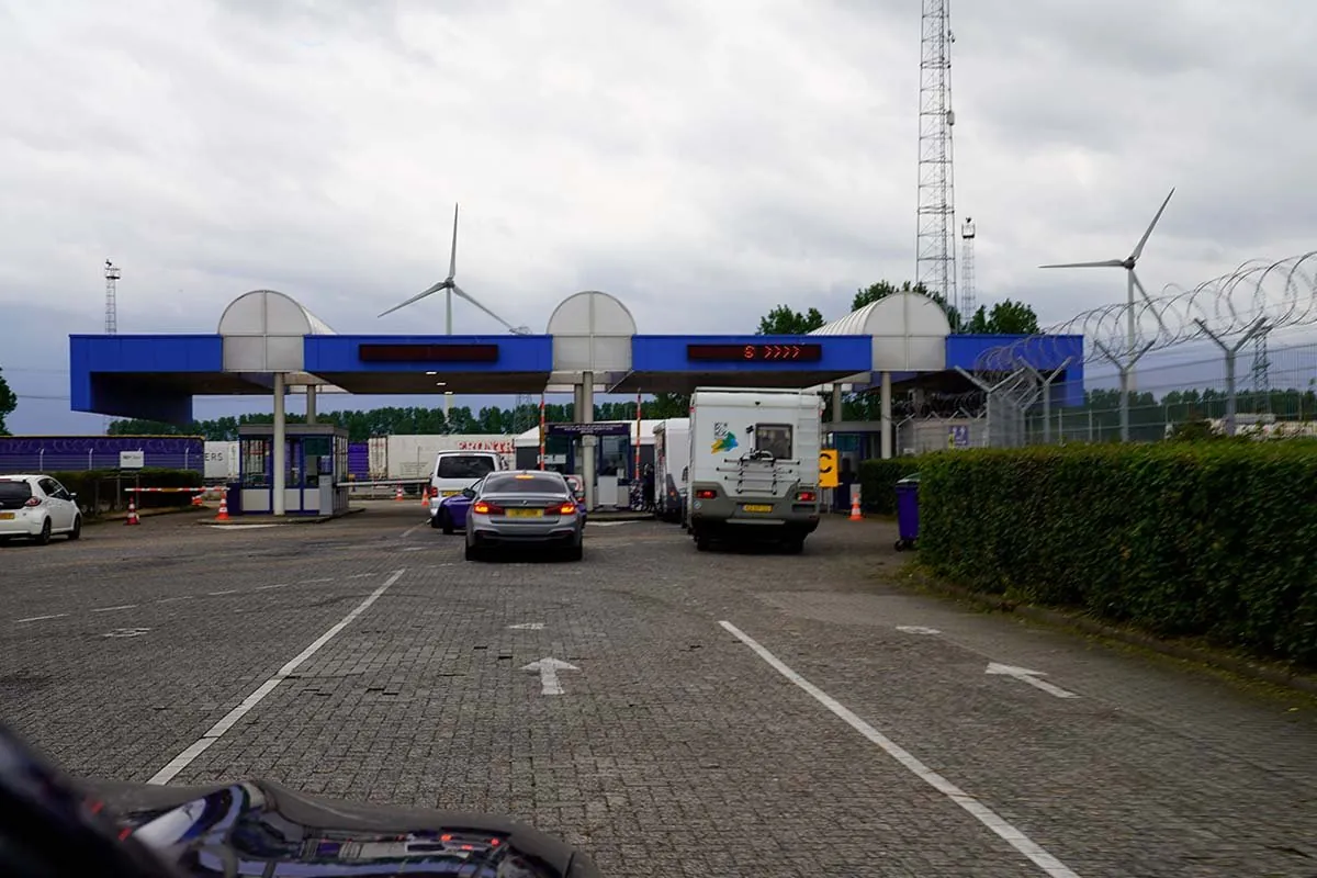 De check-in van de veerhaven: een rij cabines onder een blauwe luifel, met een auto, een bestelbus en een camper ervoor en windmolens erachter