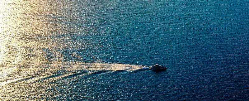 Een veerboot trekt een lang wit kielzog over een donkerblauwe zee, van bovenaf gezien bij laagstaande zon