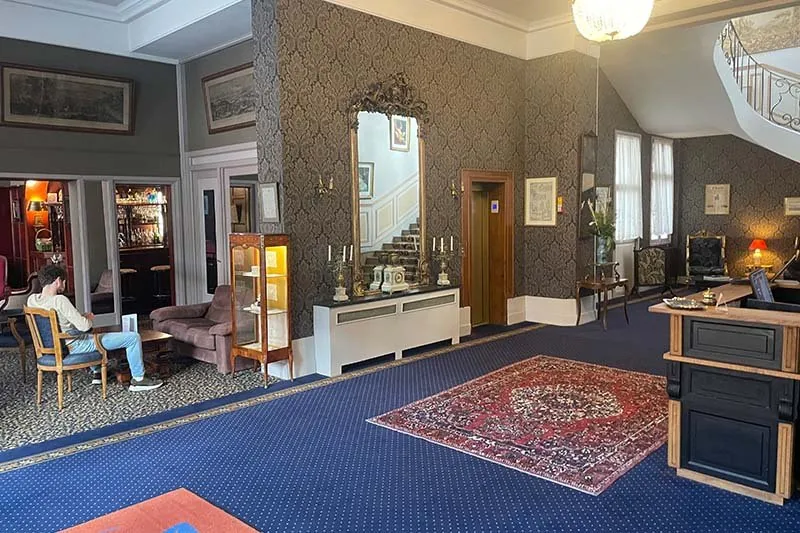 Lobby van Hotel Meurice in Calais met blauw tapijt, een vergulde spiegel boven de radiatorkast en een bar achter een openstaande deur
