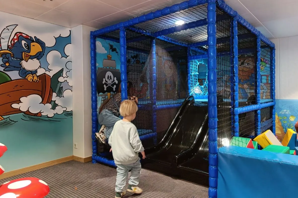 Een kind voor het blauwe klimrek met glijbaan in de Pirates Club, met een piratenschildering op de wand